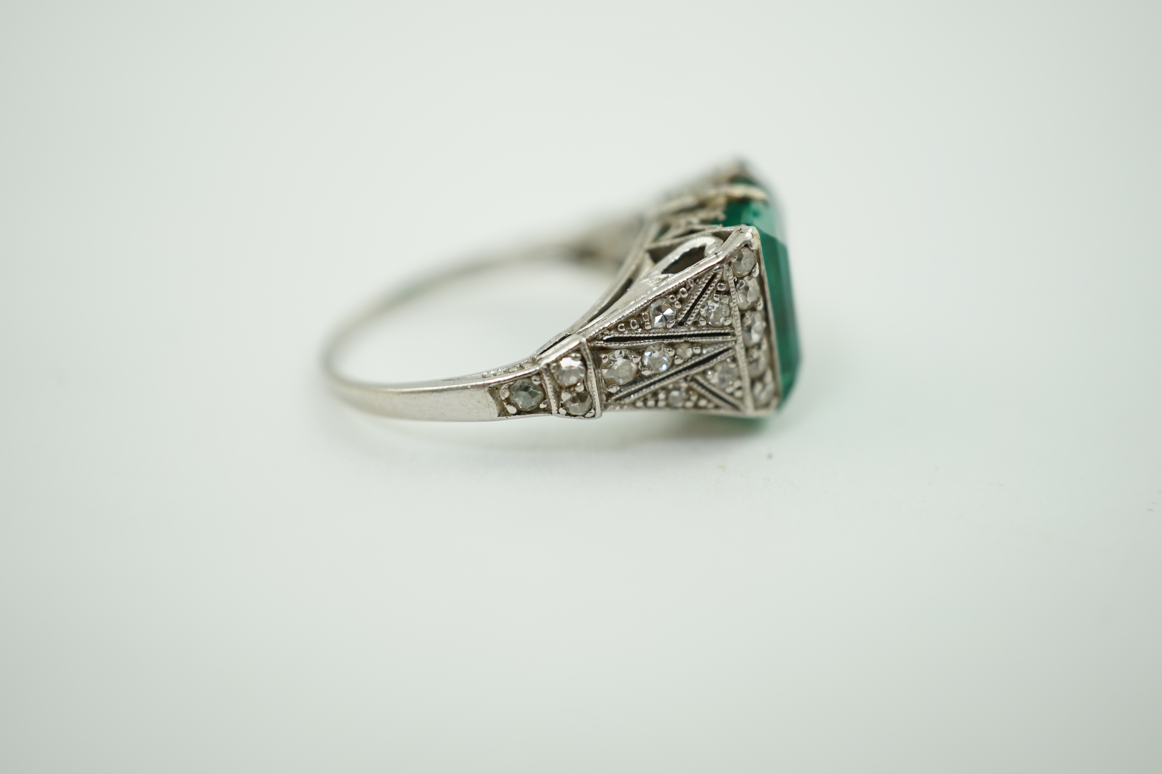 A 1920's/1930's platinum and square cut emerald set dress ring, with pierced and millegrain diamond cluster set shoulders